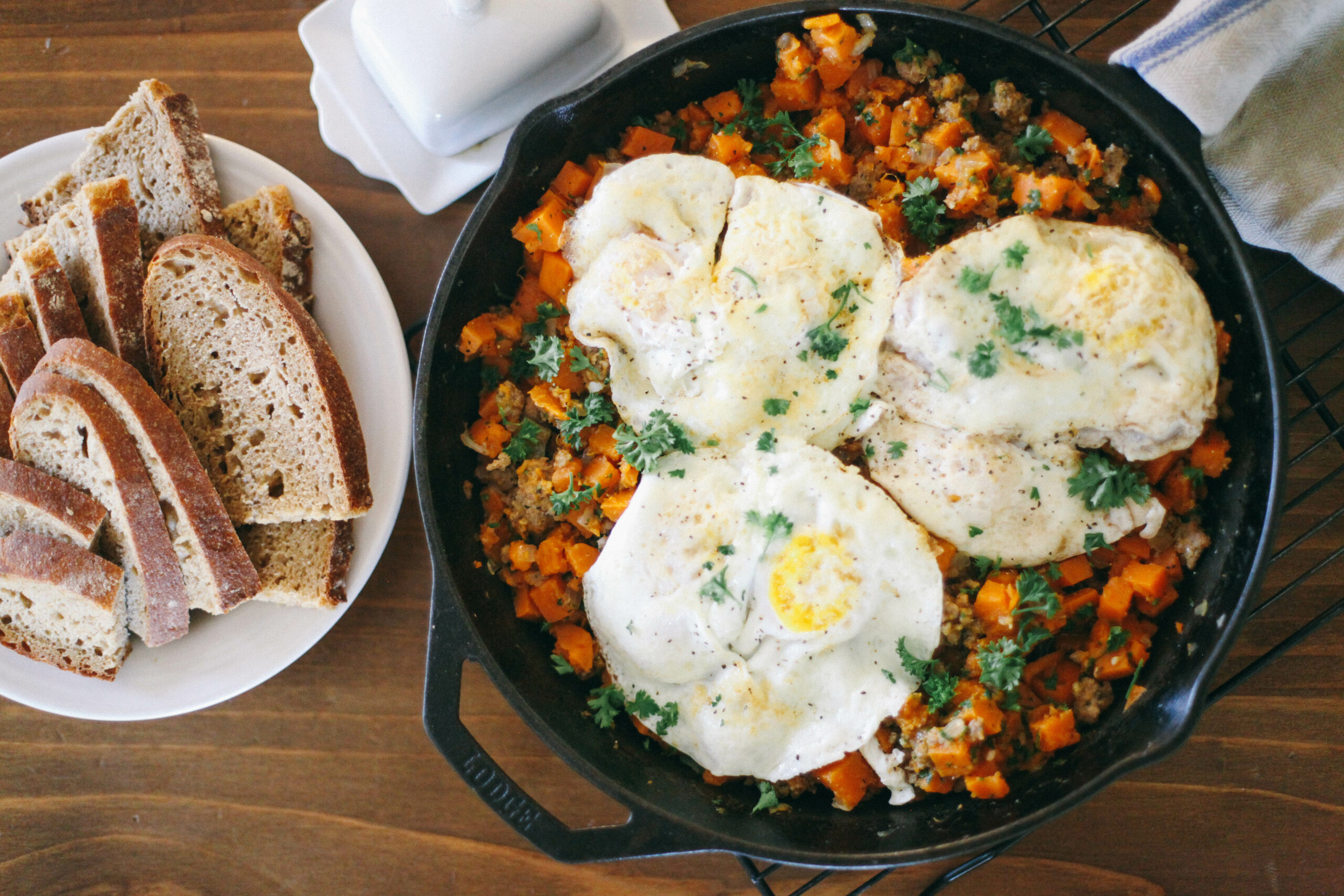 Sausage + Sweet Potato Breakfast Hash topped with fried eggs in a cast iron skillet next to slices of homemade sourdough bread