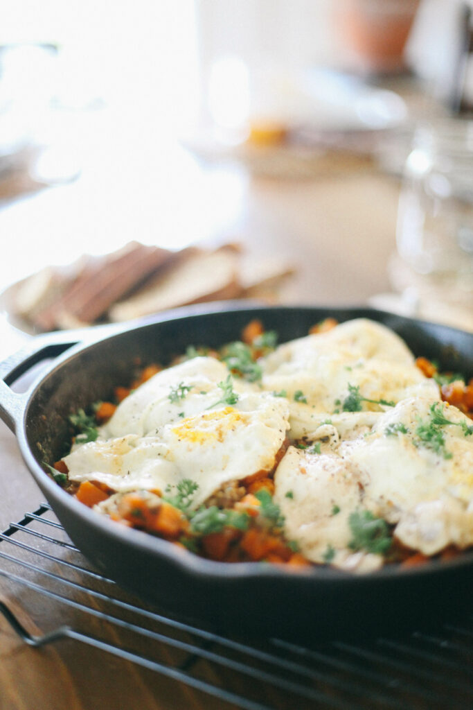 Sausage + Sweet Potato Breakfast Hash topped with fried eggs in a cast iron skillet next to slices of homemade sourdough bread