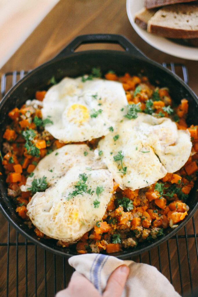 Sausage + Sweet Potato Breakfast Hash topped with fried eggs in a cast iron skillet next to slices of homemade sourdough bread