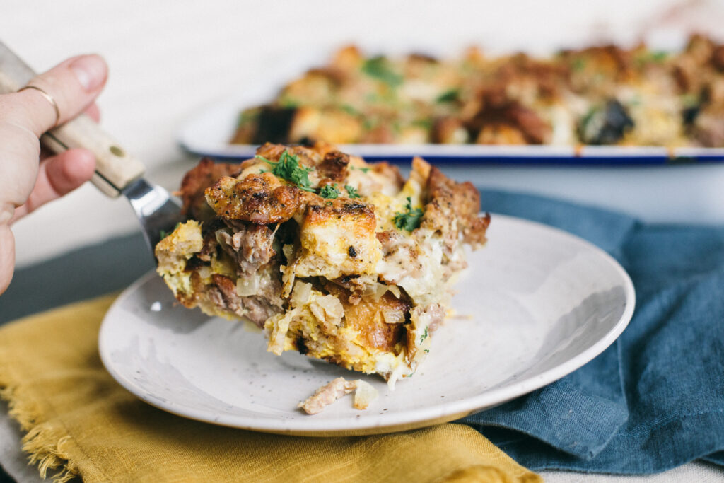 Photo of someone holding a small plate with a slice of Breakfast Strata with the full pan behind