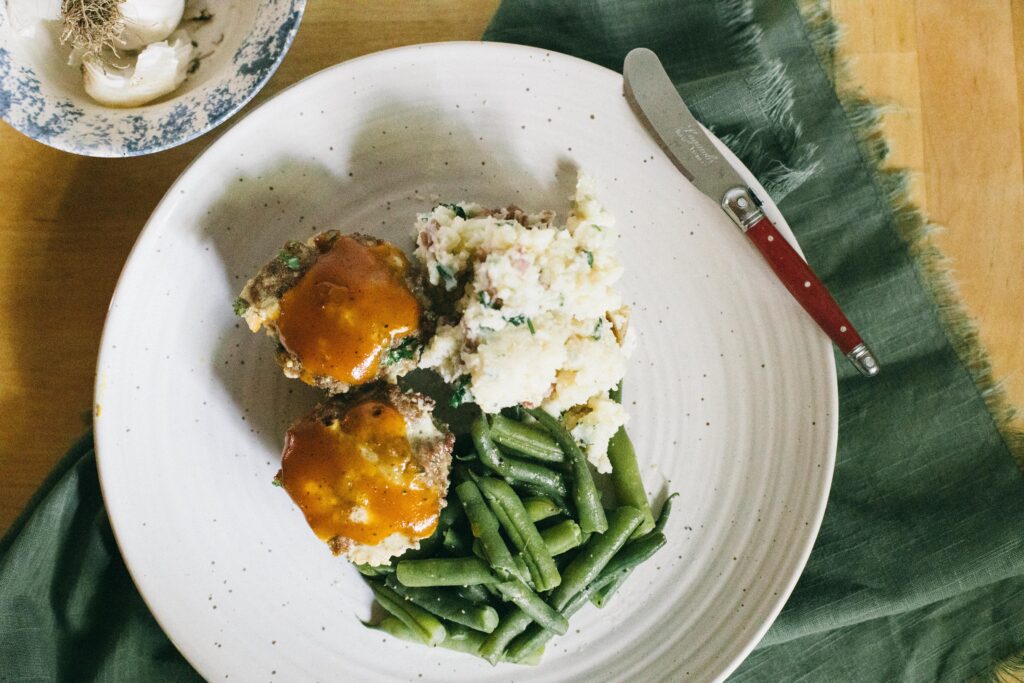 Image of a white plate with a meatloaf muffin, green beans and mashed potatoes, garden comfort food